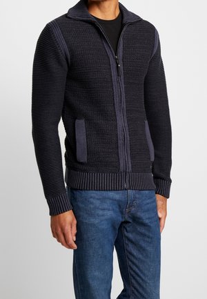 Sweater zippé en maille bleu foncé avec un col et des poignets côtelés, présentant des panneaux texturés et des poches latérales. Porté sur une chemise noire.