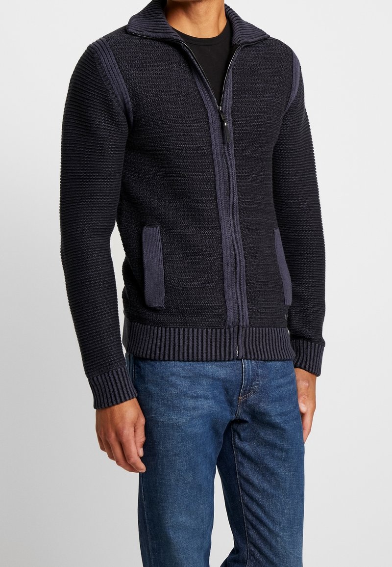 Mörkblå stickad zip-up-tröja med ribbat krage och ärmslut, med texturerade paneler och sidofickor. Bärs över en svart skjorta.