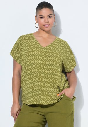 Mujer de cabello corto que lleva pendientes de aro dorados, una blusa verde con diseño de rombos y cuello en V, y pantalones verdes a juego, con una mano en el bolsillo.