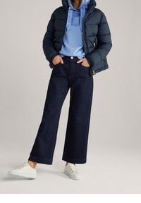 Persona che indossa una giacca a vento blu navy, un hoodie azzurro chiaro con coulisse e logo, jeans larghi scuri e sneakers bianche, in piedi con le mani nelle tasche.