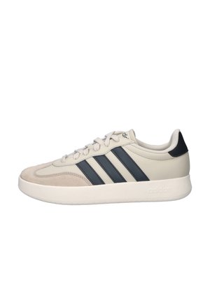 Beige Adidas sneaker met zwarte drie strepen, vetersluiting, gevoerde zool en zwarte hieltab, gezien in zijaanzicht.