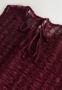 Tissu bordeaux transparent avec une texture froncée et une fermeture par lien en haut, sur fond blanc uni.