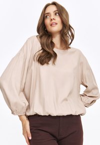 Beżowa bluzka z długim rękawem o okrągłym dekolcie i elastycznych mankietach. Miękka tkanina luźno opada w talii, tworząc swobodną sylwetkę.