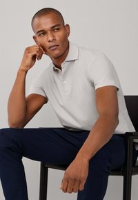 Camisa polo gris claro con cuello y tres botones, combinada con pantalones azul marino oscuro. Tela suave, mangas cortas, postura sentada.