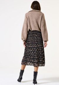 Frau mit dem Rücken in einem beigen Rippenpullover, einem schwarz-beigen gemusterten plissierten Midirock und schwarzen plateauförmigen Stiefeletten vor einem weißen Hintergrund.