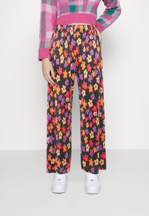 Person iført sorte vidde bukser med rød, orange, gul og lilla blomsterprint, lyserød ternet sweater og hvide sneakers.