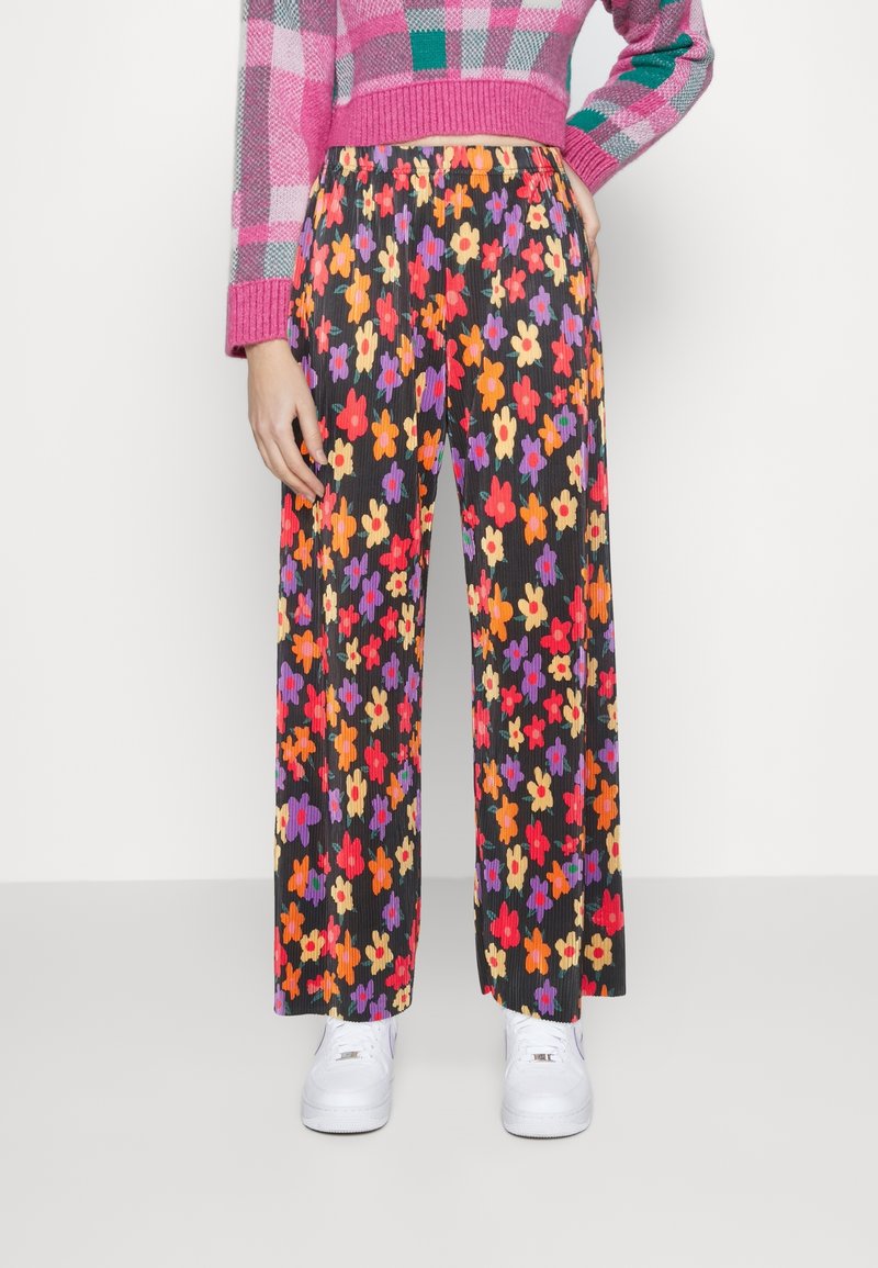 Personne portant un pantalon large noir avec un imprimé floral rouge, orange, jaune et violet, un pull rose à carreaux et des baskets blanches.