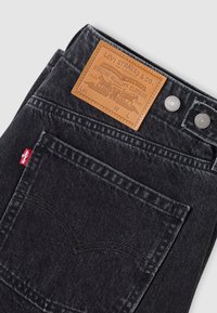 Μαύρα τζιν από denim με δερμάτινη ετικέτα που γράφει "Levi Strauss & Co." και δύο ασημένια κουμπιά. Η λευκή ραφή σχηματίζει το περίγραμμα των πίσω τσεπών.