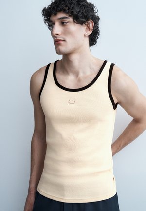 Νεαρός άνδρας με σγουρά μαύρα μαλλιά, φορώντας μπεζ αμάνικο με μαύρο περιθώριο, στέκεται με το ένα χέρι πίσω από την πλάτη του πάνω σε μια απλή ανοιχτόχρωμη φόντο.