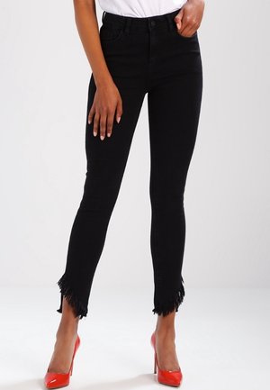 Vaqueros negros de corte ajustado con bajos asimétricos con flecos, combinados con un top blanco y zapatos de tacón rojo.