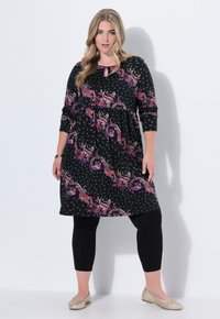 Robe noire avec des motifs paisley et floraux vifs en violet, rouge et or. Associée à des leggings noirs et des ballerines métallisées. Encolure en keyhole.
