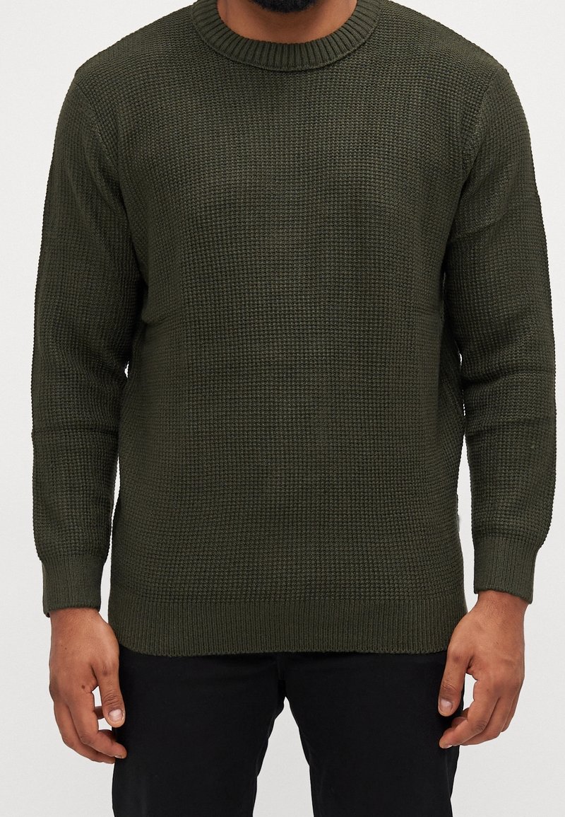 Olivgrön stickad tröja med ett strukturerat mönster, rundad halsringning och ribbade mudd och fåll, visad mot en neutral bakgrund.