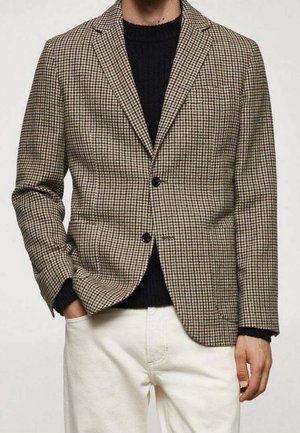 Hombre con blazer de pata de gallo marrón y beige sobre un suéter negro y pantalones blancos, con una mano en el bolsillo.