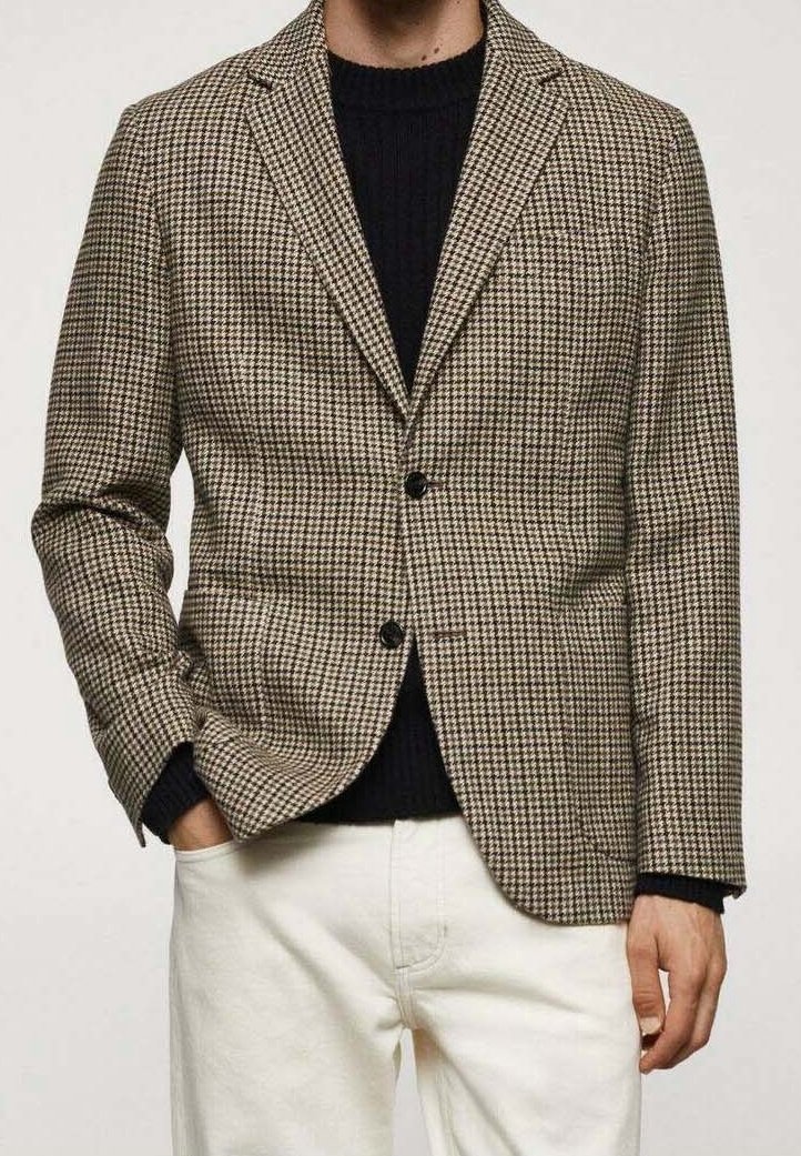 Homme portant un blazer pied-de-poule marron et beige sur un pull noir et un pantalon blanc, une main dans la poche.