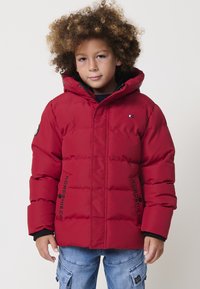 Giacca a vento rossa con cappuccio, dotata di un patch logo nero, chiusura con zip e design a righe orizzontali. Indossata sopra un maglione blu navy.