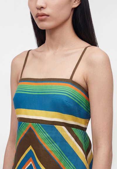 Γυναίκα που φοράει αμάνικο φόρεμα με λεπτές ράντες και πολυχρωμικές γεωμετρικές ρίγες σε μπλε, πράσινο, κίτρινο, πορτοκαλί και καφέ χρώμα.