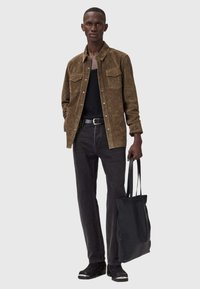 Man in een bruine suède jas, zwart tanktop, zwarte jeans, zwarte laarzen, met een zwarte gestructureerde tote bag, staand tegen een lichte achtergrond.