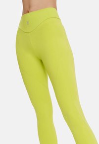 Colanți cu talie înaltă, de culoare verde lime strălucitor, realizați dintr-un material elastic cu o textură netedă; au detalii subtile asupra cusăturii.