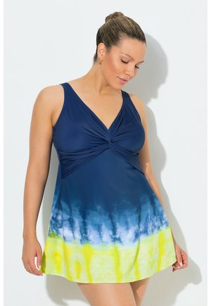 Mujer con el cabello recogido en un moño, vistiendo un vestido sin mangas de teñido anudado en azul, blanco y amarillo, mirando hacia abajo con los brazos relajados a los lados.