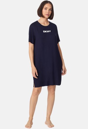 Kobieta o krótkich brązowych włosach, ubrana w luźną, granatową sukienkę na krótki rękaw z białym logo "DKNY", stojąca boso na jednolitym tle.