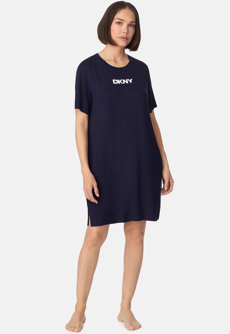 Femme aux cheveux courts bruns portant une robe bleu marine ample à manches courtes avec le logo blanc "DKNY", debout pieds nus sur un fond uni.