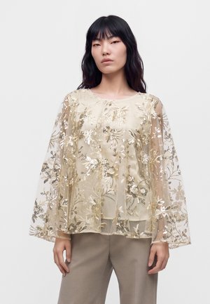 Un mannequin se tient devant un fond uni, vêtu d’un chemisier crème transparent orné de broderies florales dorées et d’un pantalon taupe.