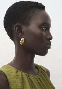 Złote kolczyki w kształcie kół zdobią profil osoby z krótką fryzurą. Osoba ma na sobie zieloną bluzkę z marszczonym dekoltem, na tle jasnym tle.