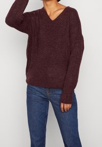 Bordeauxfärgad stickad tröja med V-ringning, avslappnad passform och långa ärmar. Tyget har en mjuk struktur och subtila ribbade detaljer. Kombineras med blå jeans.