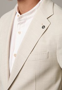 Uomo che indossa un blazer beige sopra una camicia bianca con bottoni, con una spilla a forma di croce bianca e nera sul lato sinistro del blazer.