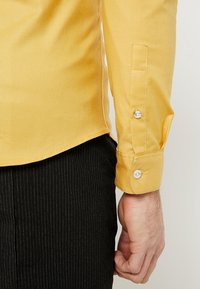 Prawa ręka w żółtej koszuli z długim rękawem z zapięciem na guzik obok czarnych spodni w prążki, ręka luźno oparta na boku.