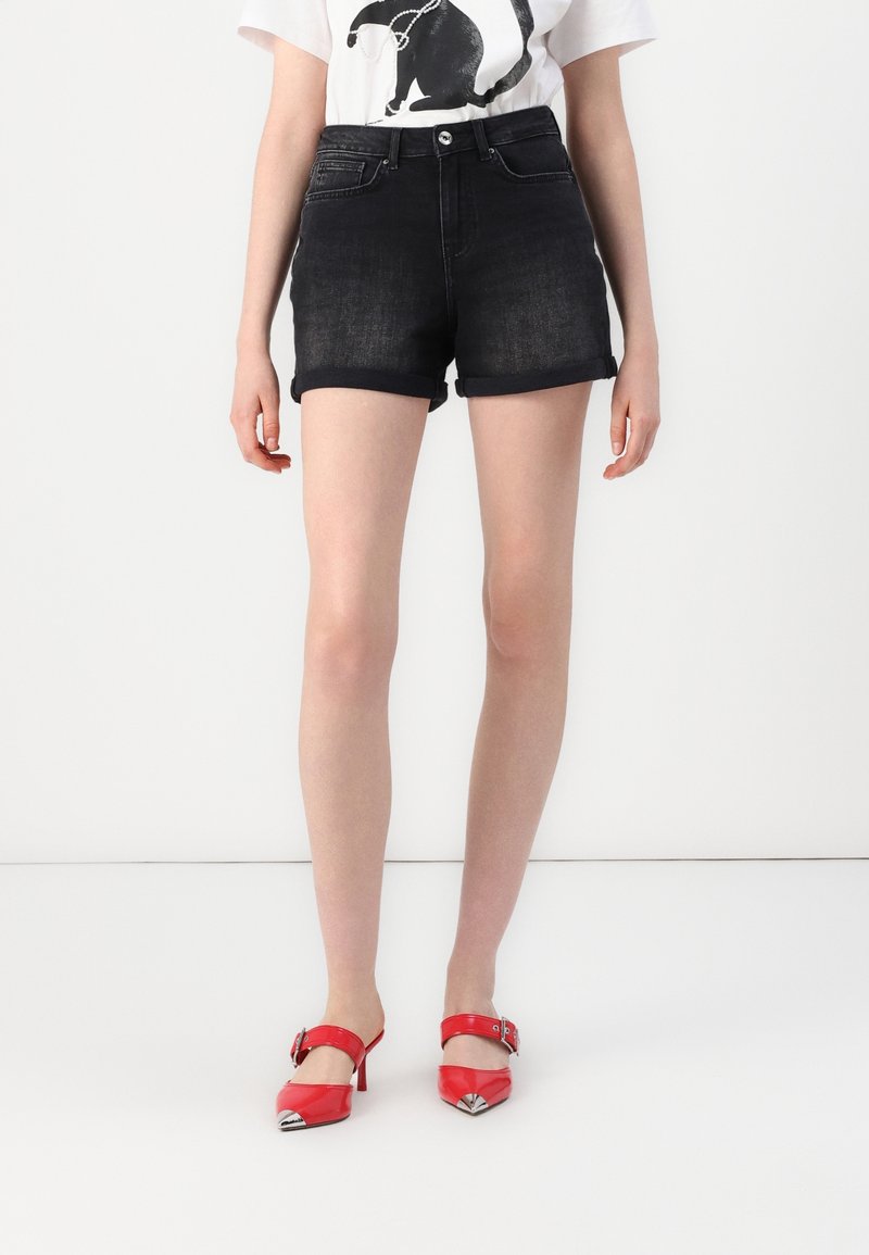 Shorts in denim neri con orli arrotolati, dotati di tasche anteriori e chiusura con bottone, abbinati a scarpe col tacco alto in vernice rossa con dettagli argentati.