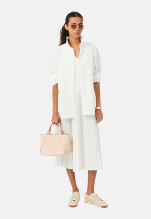 Femme aux cheveux bouclés portant une chemise blanche oversize, une jupe blanche plissée, des baskets beige, des lunettes de soleil, une montre et tenant un sac à main beige.