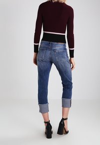 Maroon cropped neuletakki, jossa on kontrastiväriset mustat ja vaaleanpunaiset raidat, yhdistettynä käärittyihin sinisiin farkkuihin, joissa on haalistunut pesu ja takataskut.