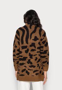 Kvinna med en brun och svart djurmönstrad tröja sedd från ryggen, i kombination med vita byxor, mot en enhetlig bakgrund.