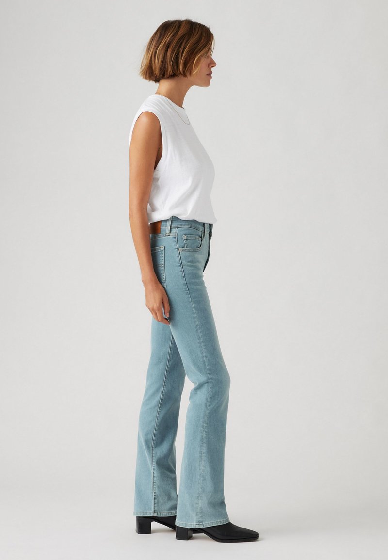 Jeans de campana azul claro con cintura alta, combinados con una camiseta sin mangas blanca. La modelo está de perfil sobre un fondo gris claro.