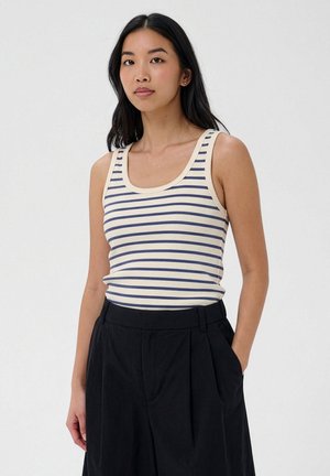 Jeune femme aux longs cheveux noirs portant un débardeur blanc à rayures horizontales bleues et un pantalon noir taille haute, debout avec une main dans la poche.