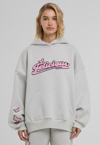 Sivi oversized hoodie s ružičastim vezom "Felicious" sprijeda, s dizajnom leptira na rukavima i kenguru džepom.
