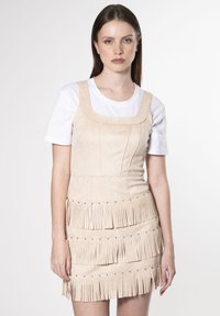 Vestido de flecos beige hecho de un material similar al ante, con escote cuadrado y flecos horizontales a lo largo del dobladillo, combinado con una camisa blanca.