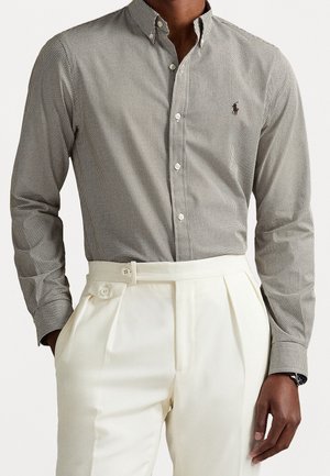 Homme portant une petite chemise à carreaux boutonnée rentrée dans un pantalon blanc taille haute avec des détails de boutons sur les côtés.