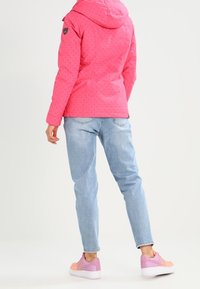 Giacca rosa a pois con cappuccio, indossata sopra jeans azzurri chiari e sneakers rosa-arancio. Presenta un logo sulla spalla.