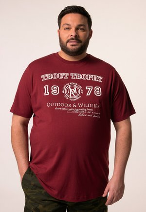 Camiseta de algodón de color marrón con texto blanco: "TROUT TROPHY 1978," con gráficos relacionados con la pesca, combinada con pantalones cortos de camuflaje.