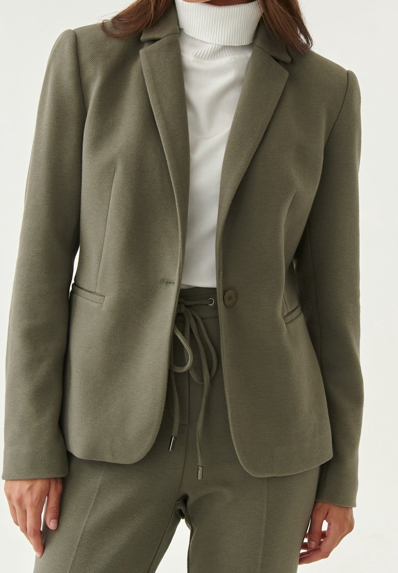 Femme portant un blazer cintré vert olive avec un pantalon assorti à taille à cordon, associé à un haut col roulé blanc.