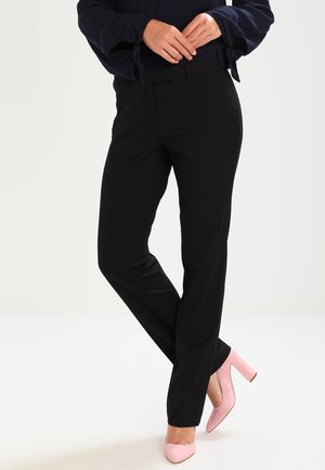 Frau trägt schwarze gerade geschnittene Hose, marineblaue Bluse und pinke High Heels, steht mit gekreuzten Beinen und verschränkten Händen.