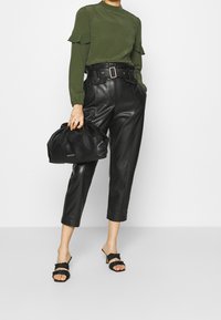 Grön långärmad topp med volanger, svarta skräddarsydda läderbyxor, svarta flätade sandaler, och en svart samlad handväska.