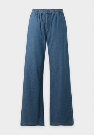 Femme debout les mains dans les poches, portant un haut à manches longues rayé noir et blanc, un jean bleu taille haute à jambes larges et des chaussures de couleur claire.