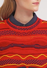 Červený pletený svetr s texturovanými vzory v žluté a modré barvě, s kulatým výstřihem a kontrastním tmavě modrým límcem pod ním.