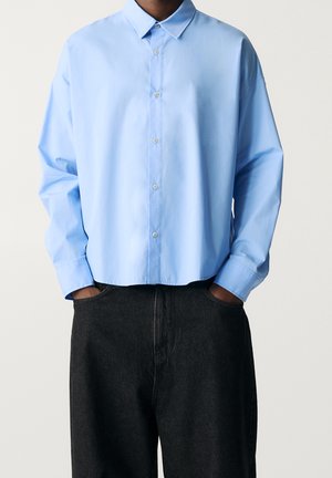 Person trägt ein locker sitzendes hellblaues Hemd mit Knöpfen und langen Ärmeln sowie schwarze Hosen, die Hände in den Vorderentaschen vergraben.