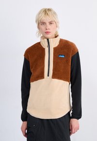 Sudadera de forro polar con una parte superior texturizada en marrón, parte inferior beige y mangas negras. Presenta un cierre frontal de media cremallera y un parche con el logo en el pecho.