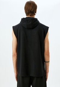 Sudadera sin mangas de color negro con una textura suave, capucha y corte relajado, que presenta agujeros de los brazos sin rematar y un dobladillo trasero ligeramente más largo.