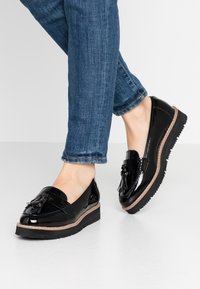 Svarta lackläder loafers med platt sula, med en dekorativ tofs och kontrasterande söm längs kanten. Bar med blå jeans.
